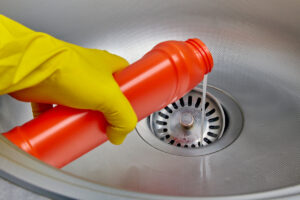A person wearing yellow dish gloves pours liquid drain cleaner into the sink