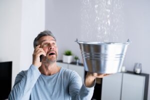 Man holding a bucket under a ceiling leak as he calls his local plumber