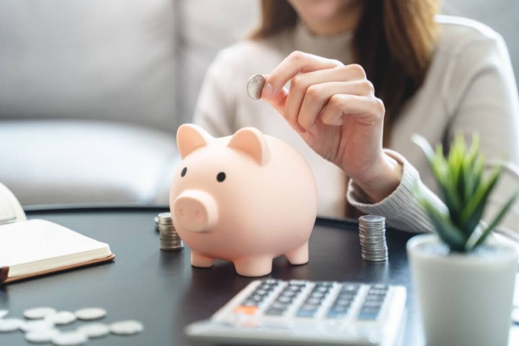 woman working on her budget with calculator and notepad - she is currently putting a penny in a piggy bank to signify saving