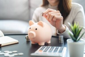 woman working on her budget with calculator and notepad - she is currently putting a penny in a piggy bank to signify saving