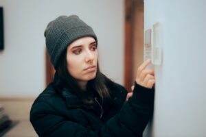woman wrapped in coat and beanie as she adjusts the thermostat because she's cold