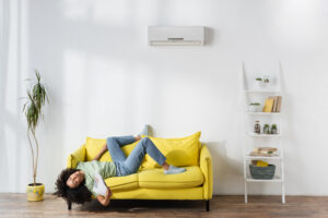 young woman sprawled out on the couch, fanning her face with a newspaper because her AC isn't cooling